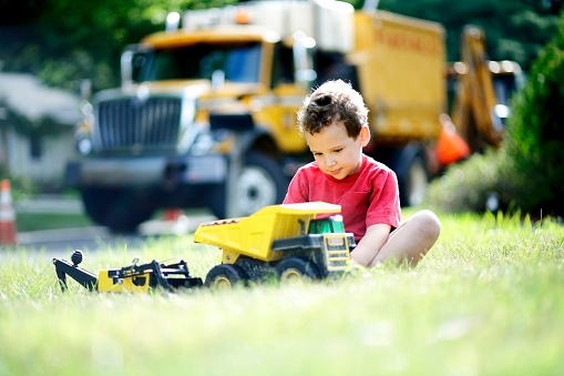 Young boy with ADHD concentrates and plays with his toy trucks on his front lawn, mimicking the roadwork taking place outside of his home on the neighborhood street.