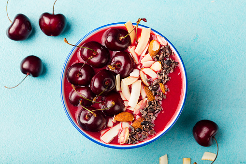 Smoothie bowl with fresh black cherries, coconut flakes, almond and cocoa nibs. Blue stone background. Top view. Copy space