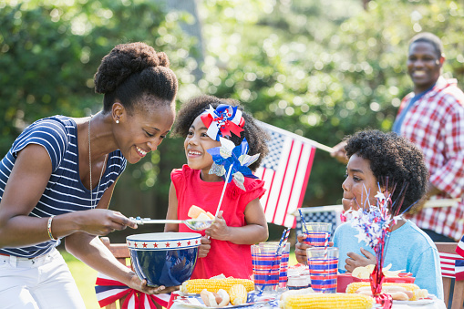 An African American mother with two mixed race children celebrating an American patriotic holiday, perhaps July 4th or Memorial Day. They are having a back yard cookout, sitting at a table decorated in red, white and blue, eating hotdogs and corn on the cob. Dad is out of focus in the background.