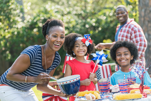 An African American mother with two mixed race children celebrating an American patriotic holiday, perhaps July 4th or Memorial Day. They are having a back yard cookout, sitting at a table decorated in red, white and blue, eating hotdogs and corn on the cob. Dad is out of focus in the background.