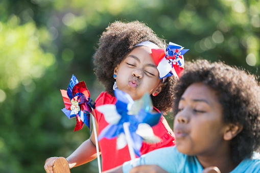 Two mixed race children having fun playing at an American patriotic event, with red, white and blue pinwheels. It could be a Memorial Day or 4th of July cookout. The focus is on the cute 7 year old little girl, eyes closed, blowing on her pinwheel. Her 12 year old brother is out of focus in the foregound. They are part black, Hispanic and Asian.