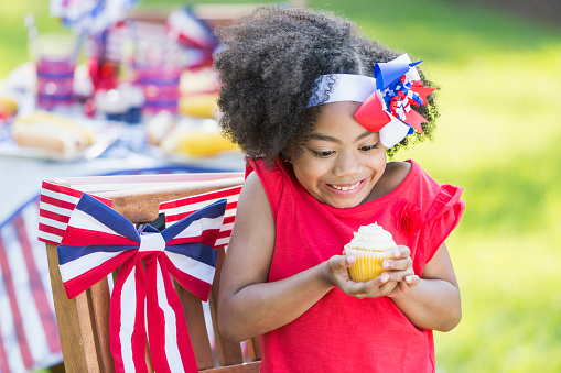 A cute little mixed race 7 year old girl enjoying a cupcake at a July 4th or Memorial Day picnic. She is part Hispanic, Asian and black. The table and chair behind her are decorated in red, white and blue, celebrating American patriotism.