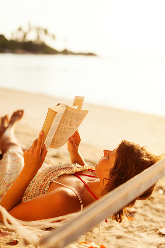 young woman reading book in hammock on a beach, sunset