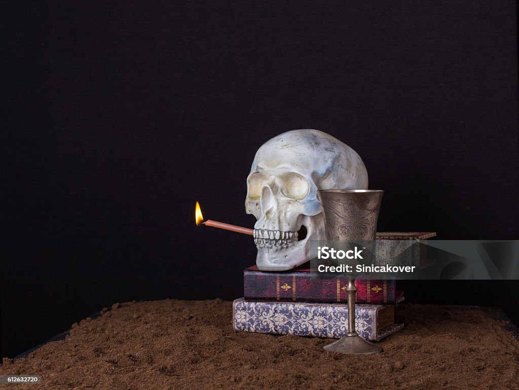 The skull is holding in his mouth a burning candle The skull on a black background standing on a pile of books and holding a lighted candle in his mouth Book Stock Photo