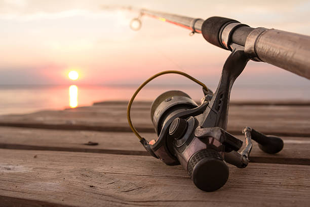 Fishing reel and rod lying on wooden pier over the Fishing reel and rod lying on wooden pier over the sunset lake. Sun over horizon fishing gear stock pictures, royalty-free photos & images