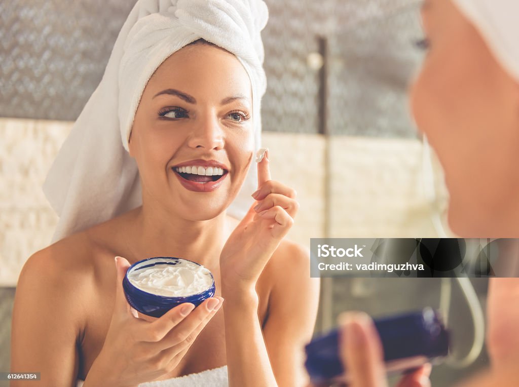 Beautiful woman in bathroom Beautiful young woman in bath towel is applying cream on her face and smiling while looking into the mirror in bathroom Moisturizer Stock Photo