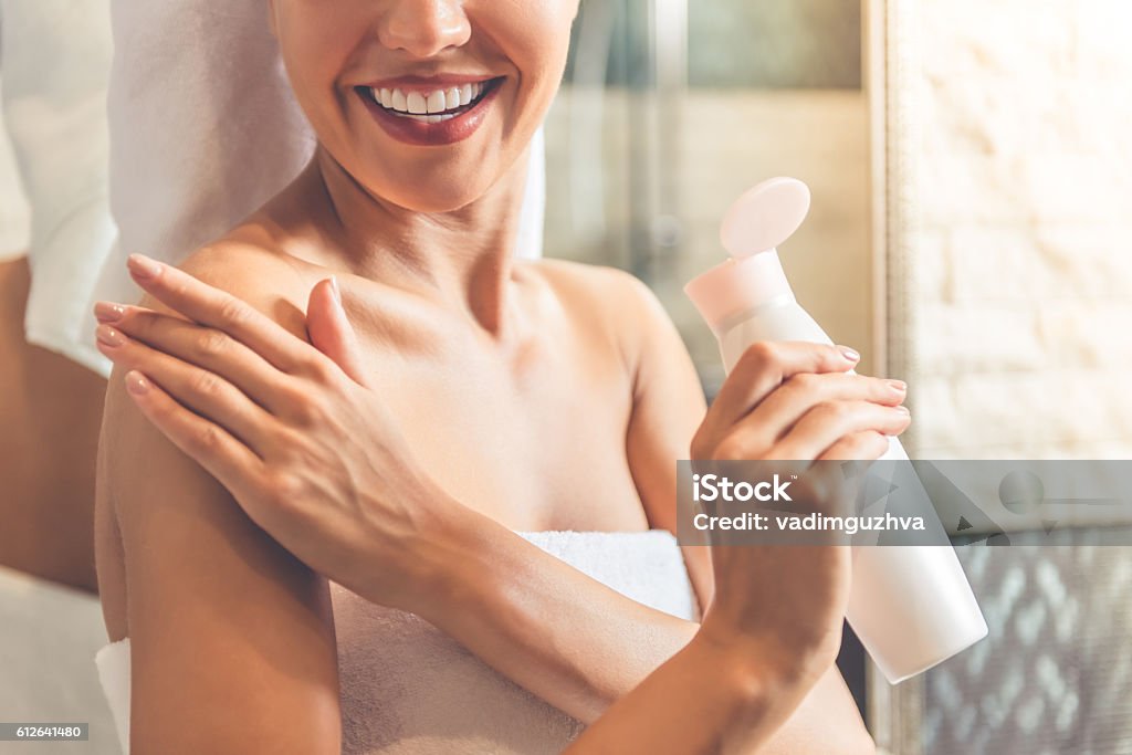 Beautiful woman in bathroom Cropped image of beautiful young woman in bath towel applying body lotion on her shoulders and smiling while standing in bathroom Moisturizer Stock Photo