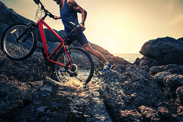 Man carrying a bike on the rock through water during sunset