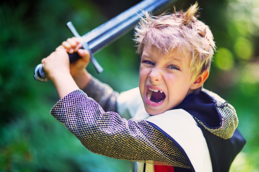 Little knight attacking with sword. Little boy dressed up as a knight is attacking imaginary foes in the forest. The boy is wielding a sword and is shouting his battle cry.