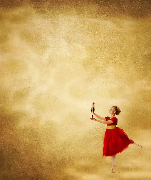 Smiling girl in festive red party dress with glittery gold ribbon holds a Nutcracker doll as she stands en pointe in ballet pose on a gold textured background with copy space for Christmas card greeting