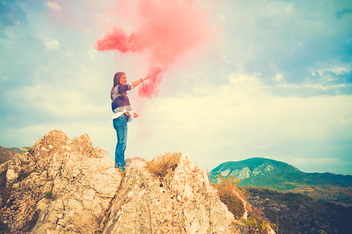 Young Hippie girl standing on a mountain rock alone, holding a torch with smoke around her.