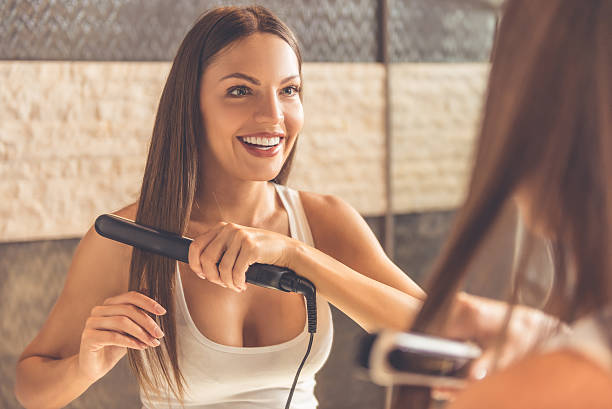 Beautiful woman in bathroom Beautiful young woman in white undershirt is using a hair straightener and smiling while looking into the mirror in bathroom chemical straightener stock pictures, royalty-free photos & images