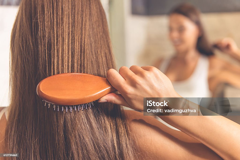 Beautiful woman in bathroom Back view of beautiful young woman in white undershirt combing her hair and smiling while looking into the mirror in bathroom Hairbrush Stock Photo