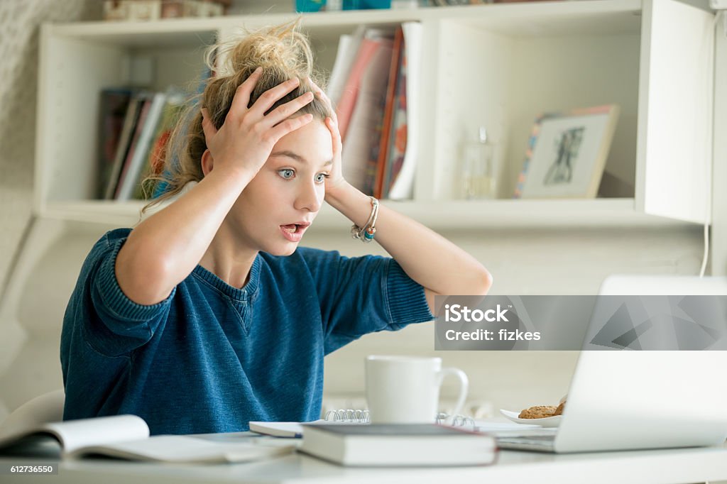 Portrait of an attractive woman at table grabbing her head Portrait of an attractive woman at the table with cup and laptop, book, notebook on it, grabbing her head. Bookshelf at the background, concept photo Women Stock Photo