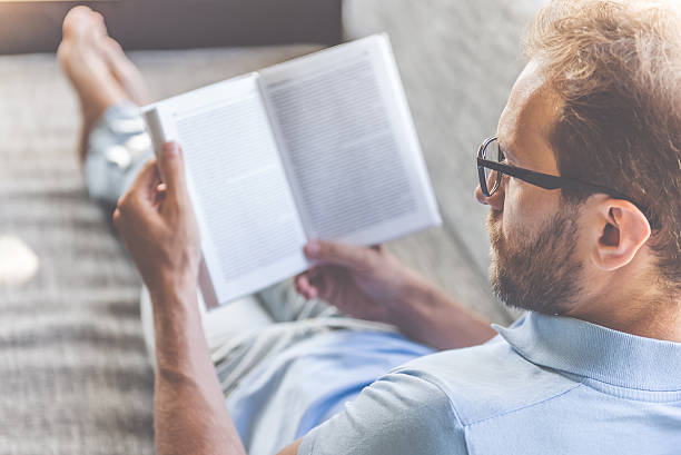 Handsome businessman at home Back view of handsome young businessman in casual clothes and eyeglasses reading a book while lying on couch at home reading glasses stock pictures, royalty-free photos & images