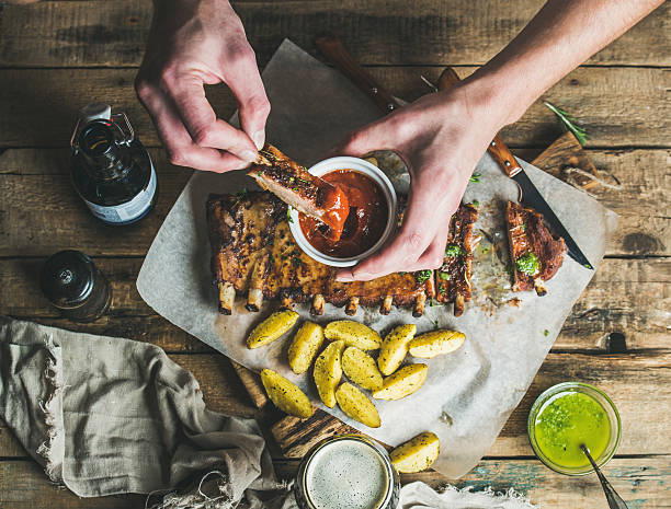 Man' s hands dipping piece of roasted pork to ketchup Man eating roasted pork ribs with potato pieces, garlic, rosemary and green herb sauce on rustic wooden table. Man's hands dipping piece of meat to ketchup in white bowl , top view rib food stock pictures, royalty-free photos & images