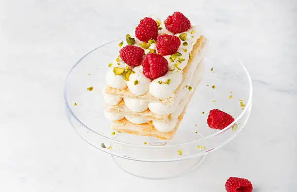 Millefeuille, Mille Feuille cake with fresh raspberries and pistachio nuts on a transparent cake stand on a white background. Copy space