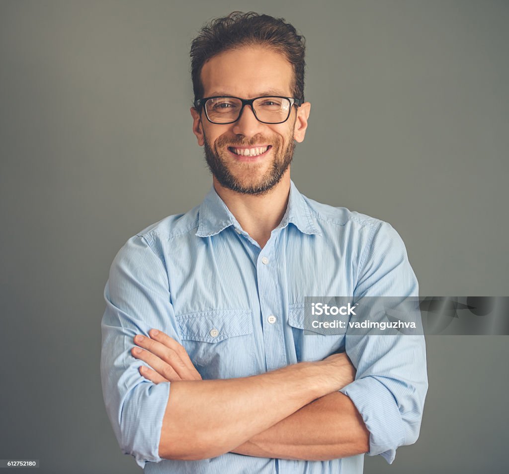 Handsome young man Handsome young businessman in shirt and eyeglasses is looking at camera and smiling while standing with crossed arms on gray background Men Stock Photo