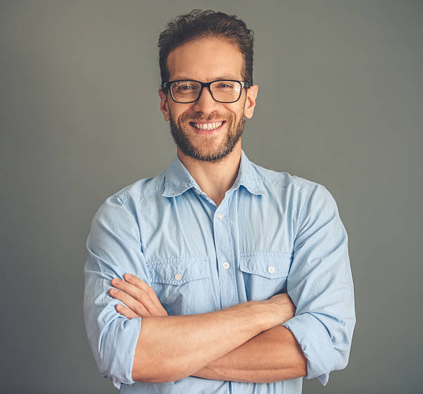 Handsome young man Handsome young businessman in shirt and eyeglasses is looking at camera and smiling while standing with crossed arms on gray background shirt photos stock pictures, royalty-free photos & images
