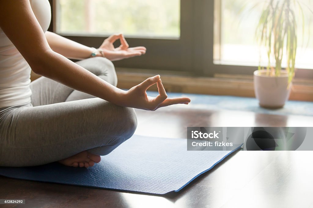 Yoga at home: meditating close-up Attractive young woman working out at home, doing yoga exercise on blue mat, sitting in Easy (Decent, Pleasant Posture), meditating, breathing, relaxing. Body close-up, focus on hand Yoga Stock Photo