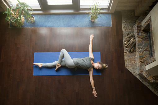 Attractive young woman working out in living room, doing yoga exercise on wooden floor, lying in Reclining Spinal Twist, Jathara Parivartanasana, resting after practice, full length, top view