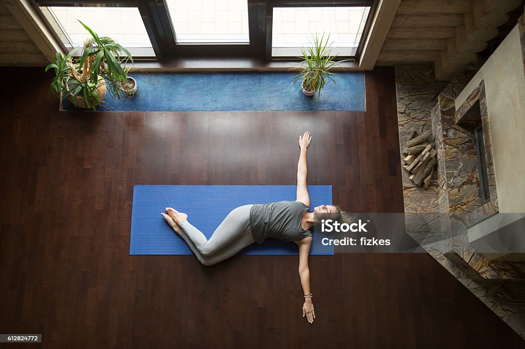 Yoga at home: Belly Twist Pose Attractive young woman working out in living room, doing yoga exercise on wooden floor, lying in Belly Twist Pose, Jathara Parivartanasana, resting after practice, full length, top view Twisted Stock Photo