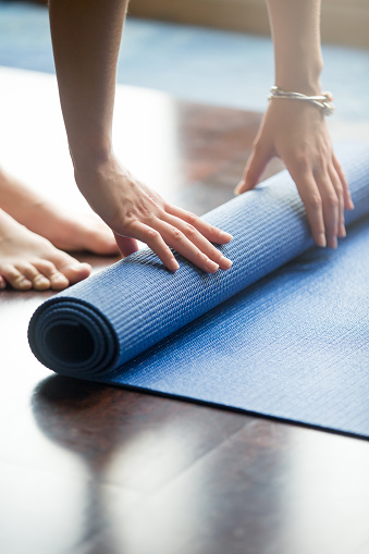 Close-up of attractive young woman folding blue yoga or fitness mat after working out at home in living room. Healthy life, keep fit concepts. Vertical image