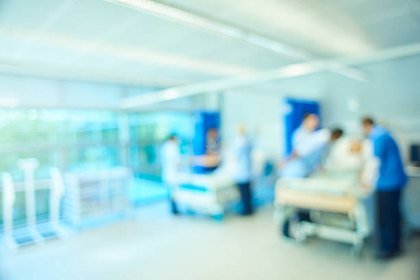 A  senior female nursing sister demonstrates the the various equipment on the training ward whilst a male staff nurse shows the medical mannequin to another group of medical student nurses . They are all standing around the hospital beds . The whole scene is defocussed to be used as a background