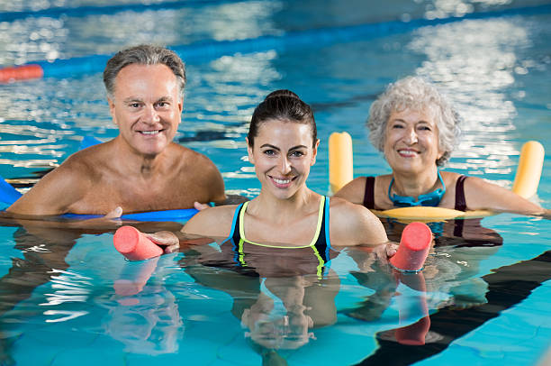 Aqua gym class Happy mature man and old woman doing aqua aerobics with foam rollers in swimming pool. Senior couple smiling with swim noodles doing aqua fitness. Smiling young trainer with mature class doing aqua gym fitness. hydrotherapy stock pictures, royalty-free photos & images