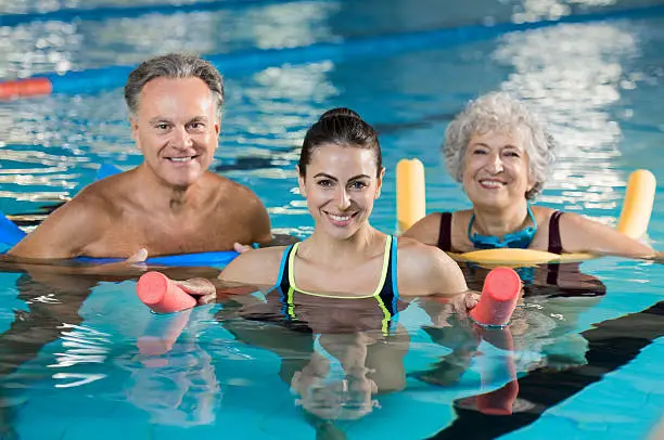 Happy mature man and old woman doing aqua aerobics with foam rollers in swimming pool. Senior couple smiling with swim noodles doing aqua fitness. Smiling young trainer with mature class doing aqua gym fitness.
