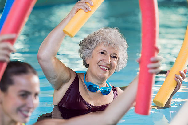 Senior woman doing aqua aerobic Smiling senior woman doing aqua fitness with swim noodles. Happy mature healthy woman taking fitness classes in aqua aerobics. Healthy old woman holding swim noodles in hand doing aqua gym with young trainer. water sport stock pictures, royalty-free photos & images