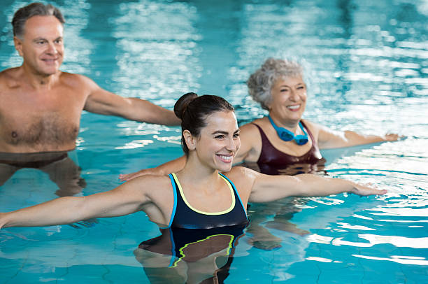 People exercising in pool Smiling fitness class doing aqua aerobics in swimming pool. Smiling young woman with senior couple stretching arms in swimming pool while doing aqua aerobics. Fit mature man and old woman exercising in swimming pool with young woman. one piece swimsuit photos stock pictures, royalty-free photos & images