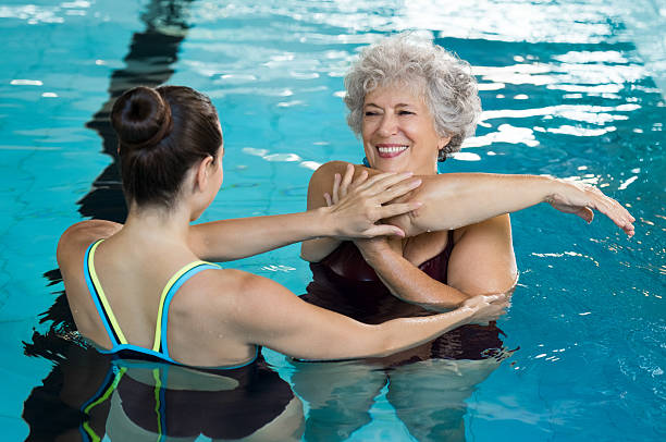 Senior woman stretching in pool Young trainer helping senior woman in aqua aerobics. Senior retired woman staying fit by aqua aerobics in swimming pool. Happy old woman stretching in swimming pool with young trainer. one piece swimsuit photos stock pictures, royalty-free photos & images