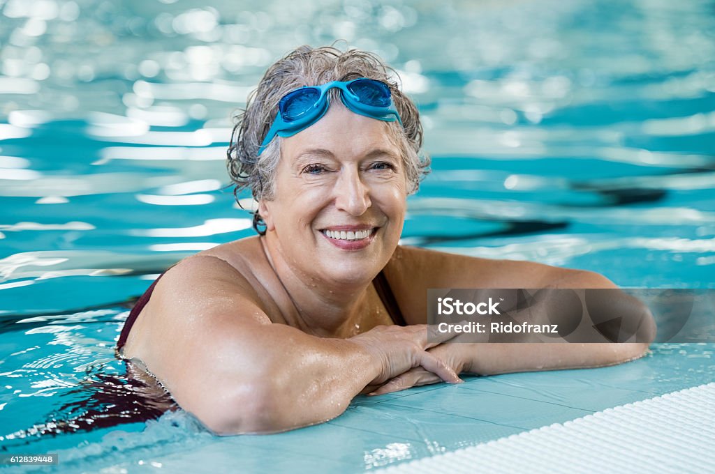 Elderly woman in pool Mature woman wearing swim goggles at swimming pool. Fit active senior woman enjoying retirement standing in swimming pool and looking at camera. Happy senior healthy old woman enjoying active lifestyle. Senior Adult Stock Photo