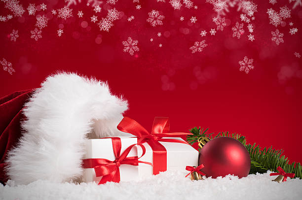 Christmas time! Christmas still life of santa claus hat, boxes with red ribbons and xmas ball. Christmas background with decorations and gift boxes on white snow over a red background. fake snow stock pictures, royalty-free photos & images