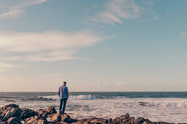 istock man standing on rocks on the shoreline looking out. 612854448