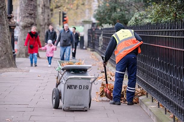 Autumn in London London, UK - December 6, 2015: Autumn i London. A street cleaner (or street sweeper) is cleaning the sidewalk (or pavement) at the Victoria Embankment in Westminster. The worker is removing red and orange coloured wilted fallen leaved from tall trees as a family (probably tourists) are walking towards him in the background. street sweeper stock pictures, royalty-free photos & images