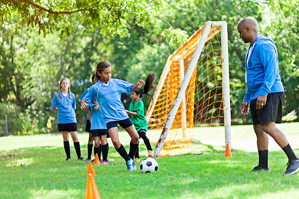 Female soccer athlete participates in practice drills Confident African American teenage soccer player runs while kicking the soccer ball around orange practice cones. Her African American male coach encourages her. Her teammates are lined up in the background. The soccer goal is in the background as well. teen goalie stock pictures, royalty-free photos & images