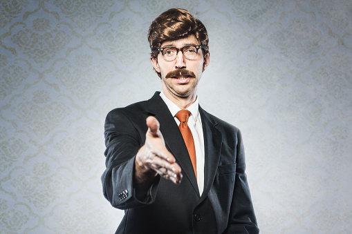 A stereotypical corporate manager type business man in full suit, big combover hairstyle and mustache looks at the camera with arm extended for a handshake.  Intentional cliche look for humor.  Horizontal image with copy space.