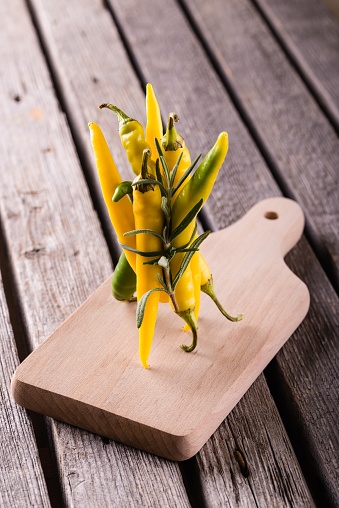 Vertical photo with bunch of green and yellow chili peppers together with sprig of rosemary herb bonded by chive. Vegetable stands on tops on chopping board and wooden grey table.
