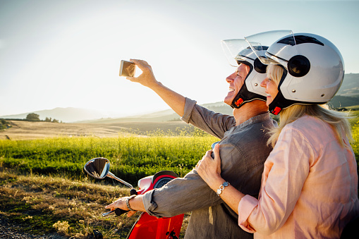 A senior couple taking a self portrait on a smart phone, they are sitting on a moped and are wearing crash helmets, ready to go off on an adventure.