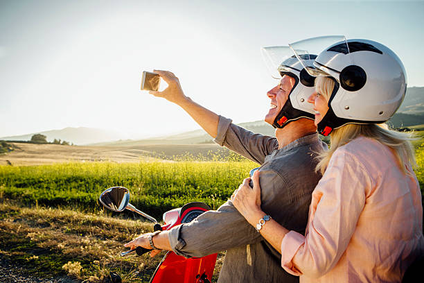 Moped Selfie A senior couple taking a self portrait on a smart phone, they are sitting on a moped and are wearing crash helmets, ready to go off on an adventure. moped stock pictures, royalty-free photos & images