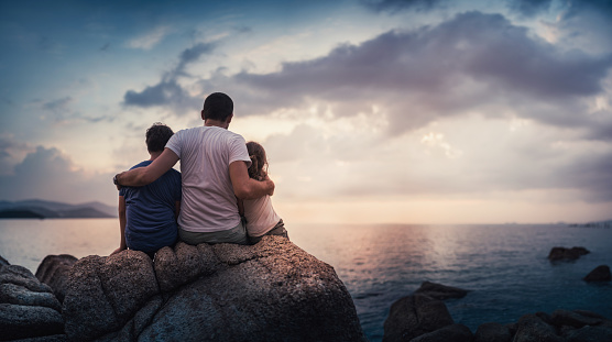 Father and his children sitting on rocks and watching the sunset over sea. Stock photo.