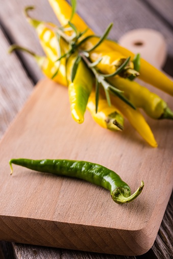 Vertical photo of single green chili pepper which is placed on wooden chopping board in front of others. Several yellow peppers in a bunch with rosemary herb bonded by chive.