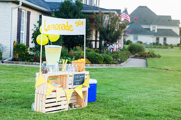 Lemonade stand set up in front yard A refreshing lemonade stand is set up in the front yard of a home in the suburbs on a sunny summer day. A beverage dispenser, basket of fresh lemons and glasses are on the stand. Balloons are attached to the stand. A banner across the top of the stand lists the price of the lemonade. images of lemonade stands stock pictures, royalty-free photos & images