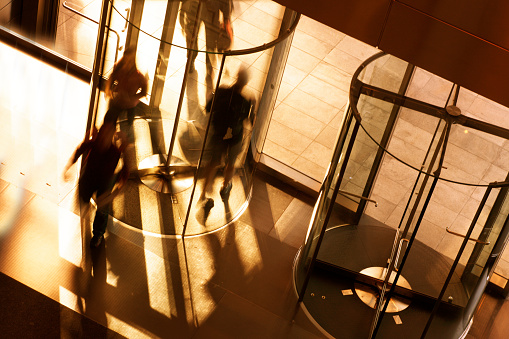 A high angle view of a people entering and exiting an office building through a revolving door. A slow shutter speed allows for the anonymity of the people as they quickly move through the rotating door.  The late afternoon sun throws the shadows of busy people and glass across the floor.