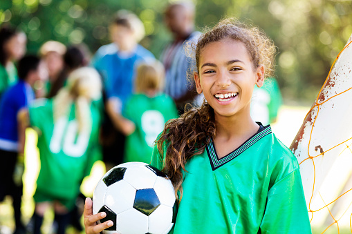 Pretty African American girl smiles proudly after winning soccer game. She is holding a soccer ball and is wearing a green jersey. She has brown curly hair back in a pony tail. Her teammates, oposing team, coach and referee are in the background along with the soccer goal.