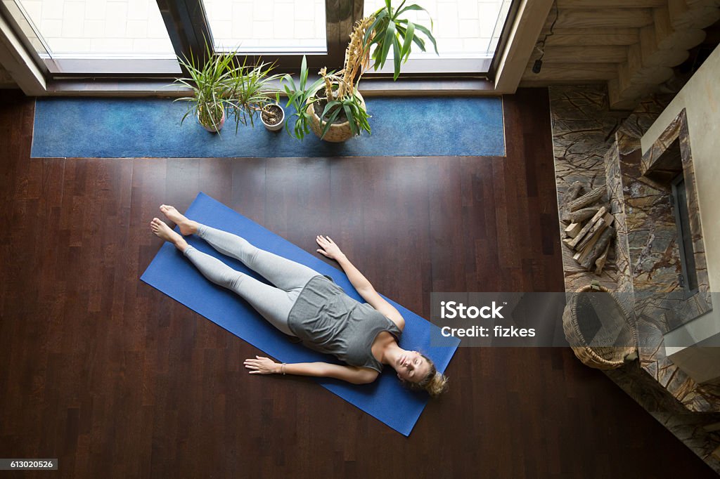 Yoga at home: relaxation Attractive young woman working out in living room, doing yoga exercise on blue mat, lying in Shavasana (Corpse, Dead Body Posture), resting after practice, meditating, breathing. Full length, top view Shavasana Stock Photo