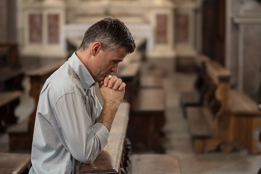 Religious man kneeling at the pew in the Church and praying with hands clasped
