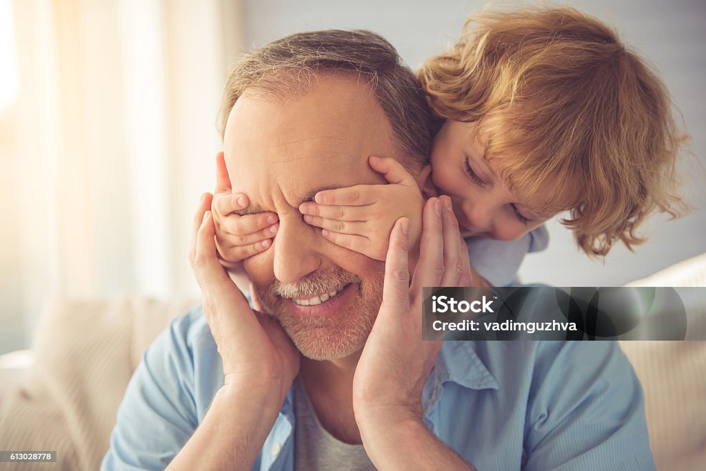 Grandpa and grandson Cute little boy is covering his grandpa's eyes and smiling while playing with him at home Grandfather Stock Photo